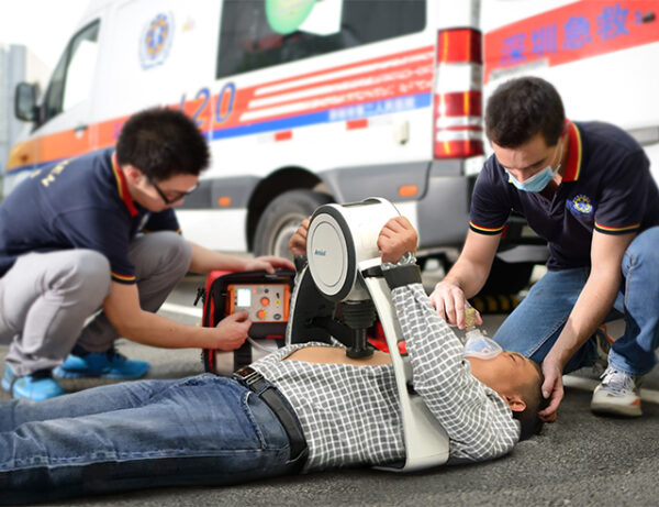 Ambulanc Amoul CPR E6 - Thoraxkompressionsgerät - zwei Rettungsdienst-Mitarbeiter vor Krankenwagen führen eine Wiederbelebung mit automatisierter Herzdruckmassage durch.
